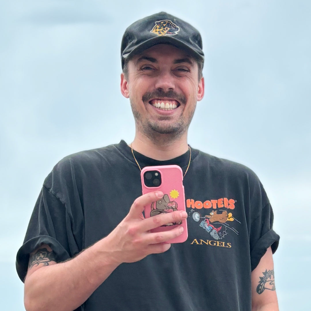 Artist Cam Miller smiles while holding the Bubblegum Pink Snakebite Pela Phone Case that he designed. He wears a black cap and a black Hooters Angels t-shirt, showcasing his casual style. The background is a cloudy sky.