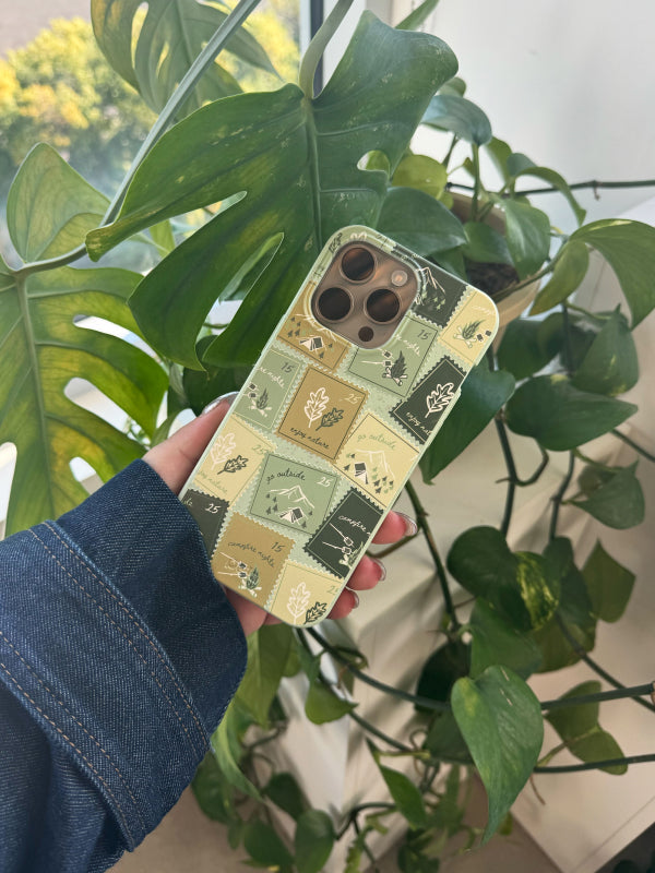 A hand holds the Pela Camp Stamps Phone Case, which features a charming design of green and beige camping-themed stamps with illustrations of tents, oak leaves, and nature icons. The case exudes a nostalgic, outdoorsy vibe, perfect for nature lovers. The background includes lush green houseplants, complementing the eco-friendly theme.