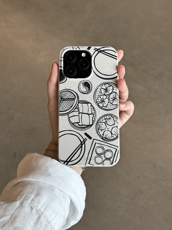 A hand holds a Pela phone case featuring the Dimsum Doodles design. The case has a speckled off-white background with black line art illustrations of dim sum dishes, including dumplings, buns, and chopsticks, arranged in circular bamboo steamer outlines. The phone has a triple-lens camera, and the person is wearing a white long-sleeve shirt. The design is minimal and food-themed, and the case is made from Pela’s compostable material.
