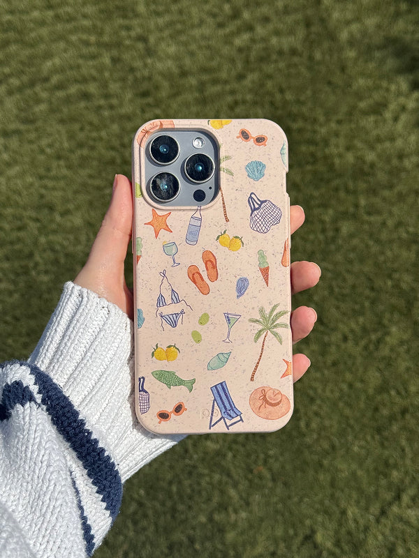 A hand holds a Pela phone case featuring the Coastline Sketch design. The case has a speckled beige-toned background decorated with colorful, hand-drawn beach icons like palm trees, flip-flops, seashells, sunglasses, cocktails, and swimsuits. The phone has a triple-lens camera, and the eco-friendly case adds a playful, summer-ready touch. The background is a grassy green surface in bright natural light.