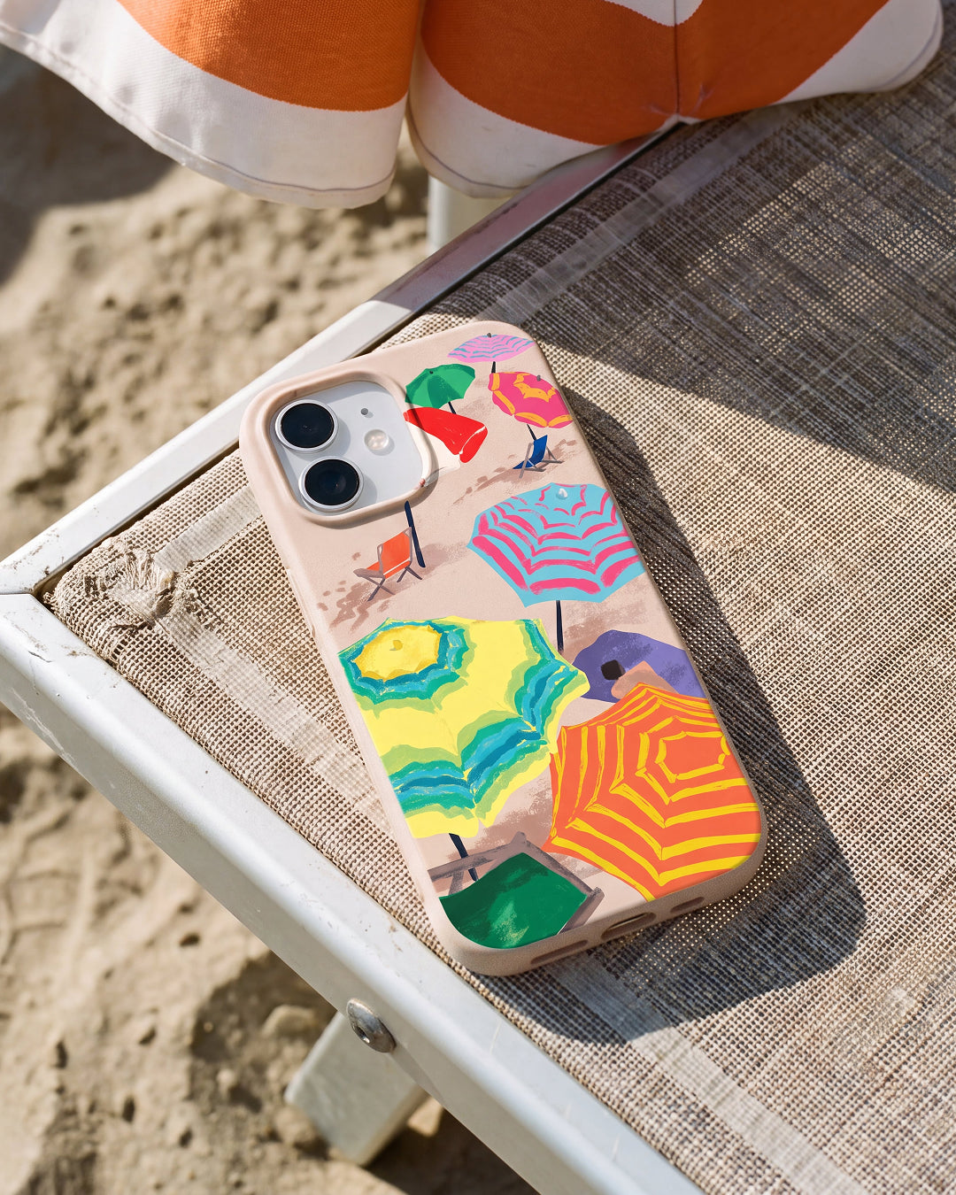 A Pela By the Beach phone case featuring a colourful illustrated aerial view of beach umbrellas in yellow, orange, green, blue, and pink, resting on a weathered white beach chair with sand and an orange and white striped beach bag visible in the background.