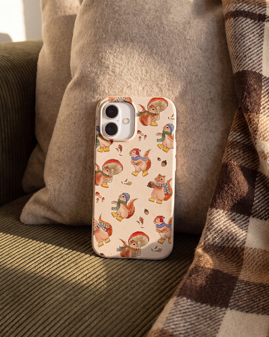 A Pela Autumn Squirrels phone case in Seashell featuring illustrated squirrels wearing cozy scarves, knit hats, and yellow rain boots while carrying mushrooms and acorns amongst falling autumn leaves, propped against a grey linen cushion on an olive green corduroy sofa with a brown plaid blanket draped beside it.