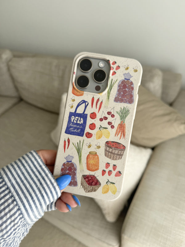 Hand holding Pela’s Market Finds Phone Case, featuring colorful illustrations of fresh produce, including carrots, strawberries, lemons, and jars of honey, inspired by farmers’ markets. The case is eco-friendly and showcases vibrant, hand-drawn designs.