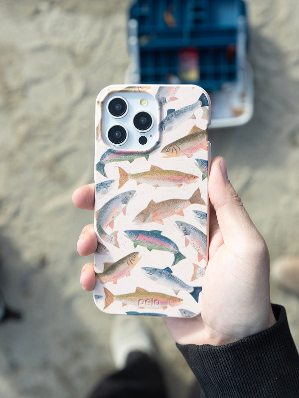 A hand holds a Pela phone case with a speckled off-white background featuring colorful rainbow trout illustrations. Made from compostable material, the case combines eco-friendly design with an outdoorsy vibe. A tackle box sits in the blurred background.