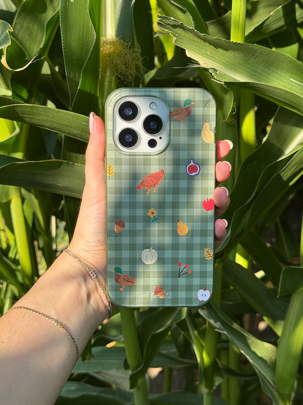 Hand holding the Harvest Folk Phone Case by Pela in front of tall green corn stalks, featuring a green gingham background with autumn icons like chickens, ducks, pumpkins, figs, acorns, apples, and leaves.