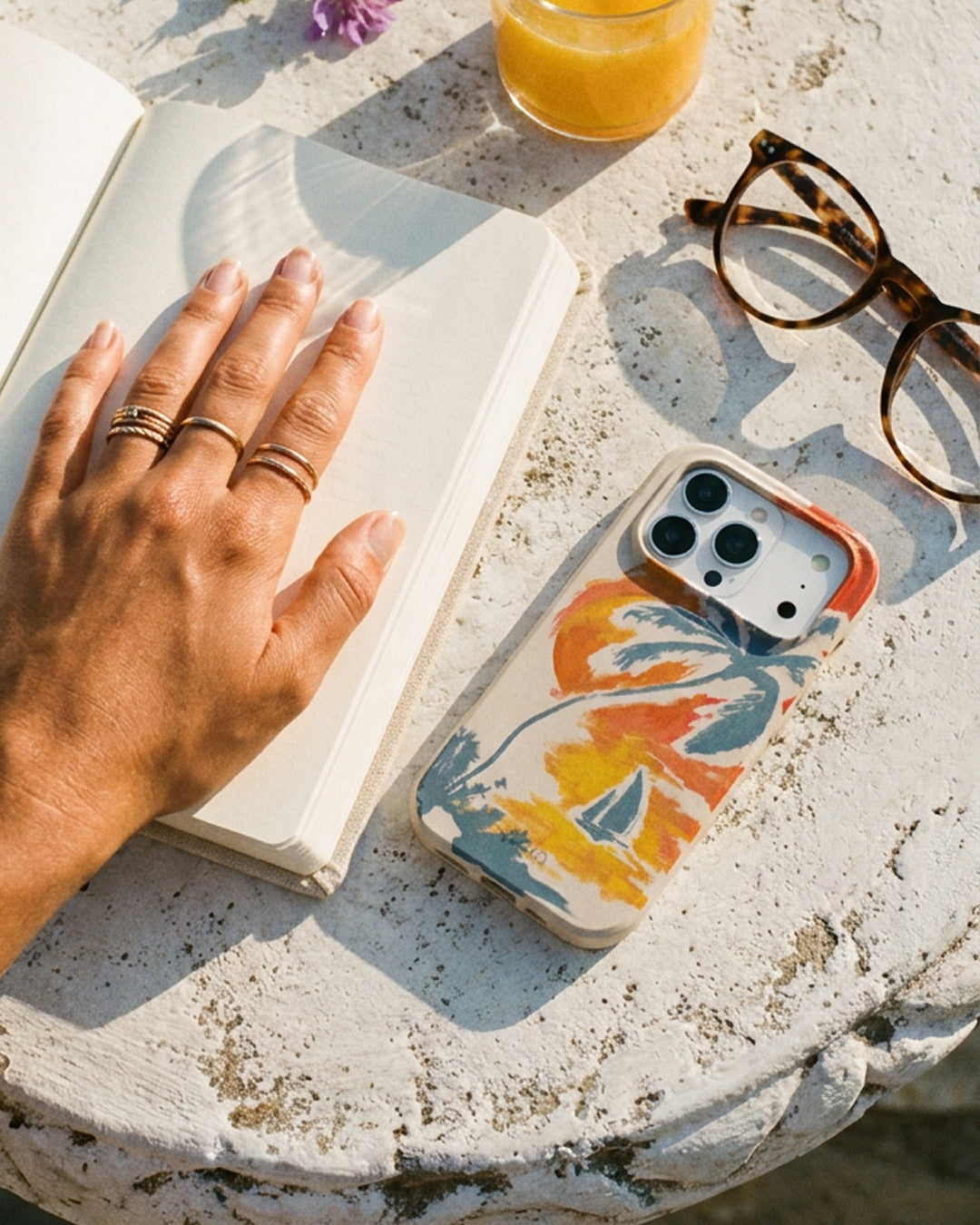 A Pela Paradise Palm phone case in warm orange, yellow, and blue brushstroke botanical print resting on a weathered stone table in bright sunlight, styled alongside a open book, tortoiseshell glasses, and a glass of orange juice — with a ringed hand resting on the book in the foreground.