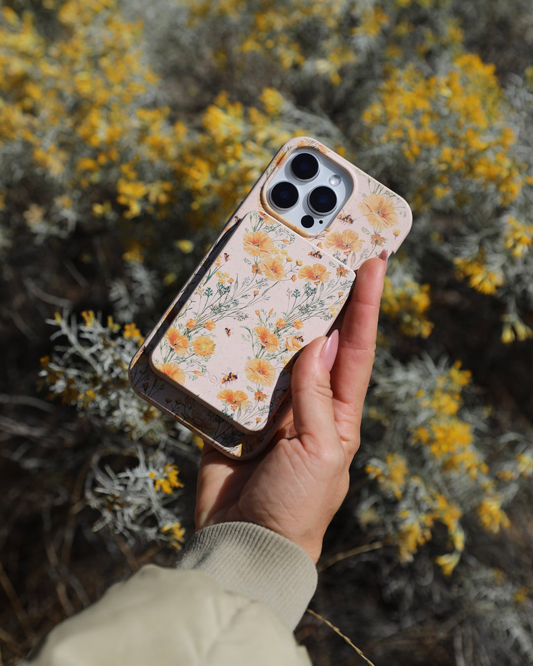 A hand holding a Pela Vintage Bee phone case with a matching Vintage Bee JuiceBox PowerBank attached, featuring delicate illustrated golden California poppies, wildflowers, and bees on a soft blush background — held up against a blurred backdrop of silver-leafed shrubs with yellow wildflowers in bright natural sunlight.
