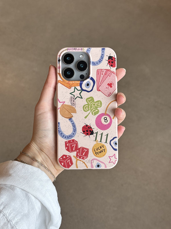 A hand holds a Pela phone case featuring the Lucky Me design. The case has a soft beige, speckled background decorated with colorful lucky charms including dice, ladybugs, four-leaf clovers, evil eyes, horseshoes, a fortune cookie, playing cards, and a “Lucky Girl” clover. The phone has a triple-lens camera, and the case is made from Pela’s compostable material, blending playful, good-luck-inspired graphics with sustainable style. The background is a neutral, softly lit surface.