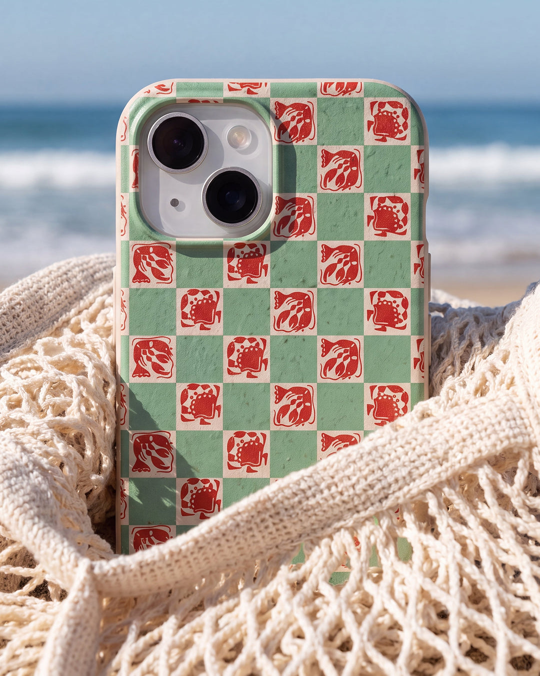 A Pela Coastal Crustaceans phone case in sage green and red checker print featuring illustrated crabs, lobsters, and fish, nestled in a cream macramé beach bag with the ocean and sandy shore blurred in the background.