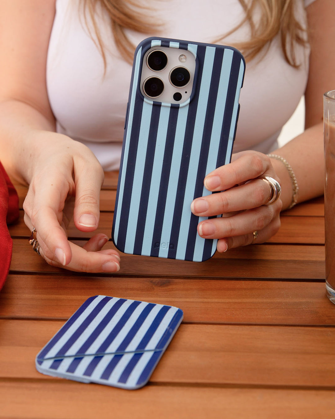 A woman holding a Pela Baby Blue Stripes phone case in navy and light blue vertical stripes, seated at a wooden outdoor table with a matching navy and white striped Pela Mini Wallet resting on the table beside her — wearing silver rings and a pearl bracelet with a glass of water in the background.