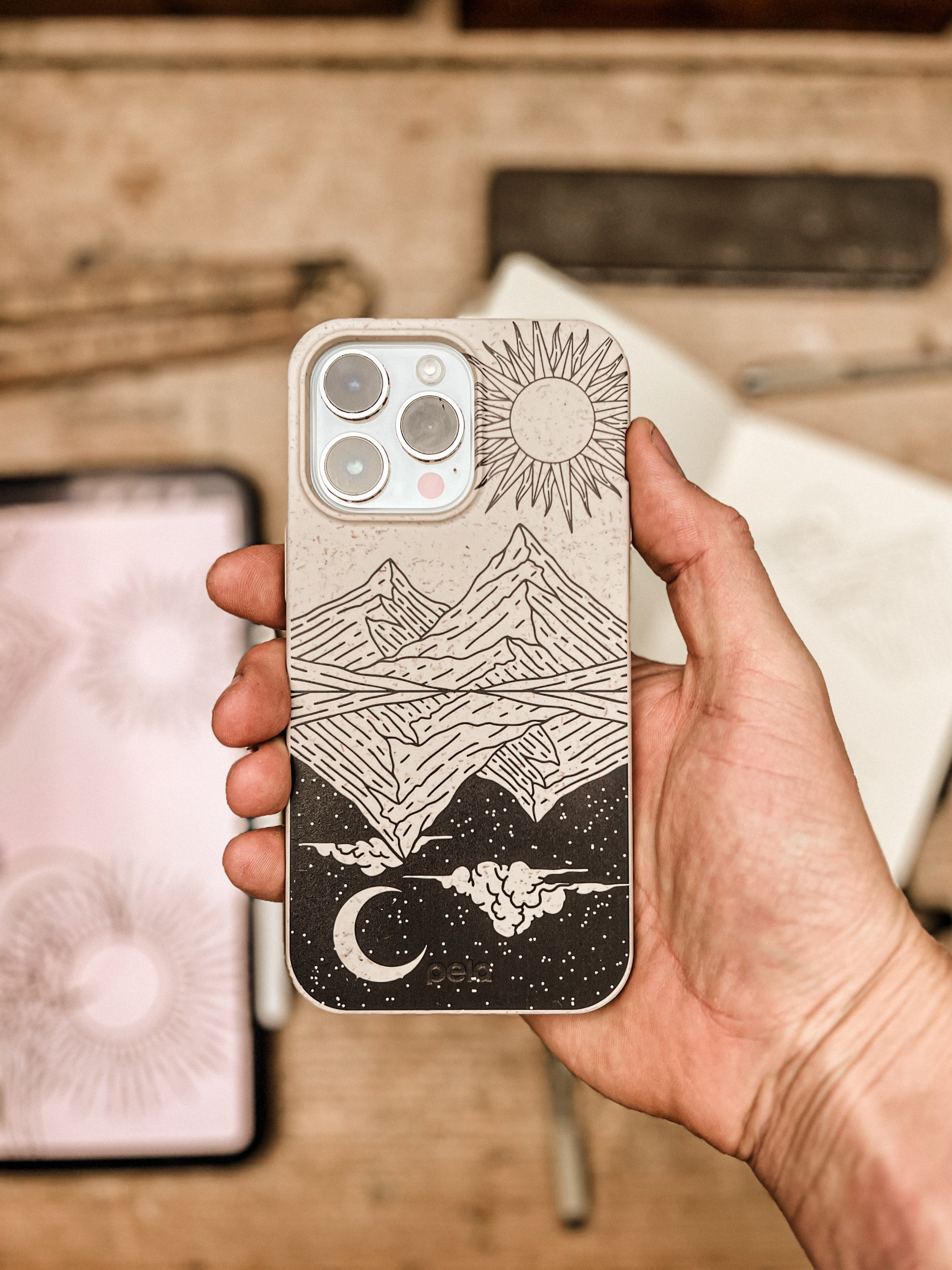 Hand holding iPhone with Pela's Horizons phone case featuring hand-drawn mountains, radiant sun above, crescent moon below, and starry night sky in intricate black line art on cream background from the Ollie Smither Artist Series Collection