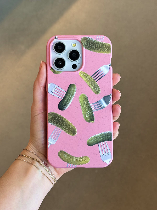 A hand holds a Pela phone case with a playful Pickle Party design. The case features a speckled pink background with realistic green pickles and silver forks scattered across it. The bold contrast between the bright pickles and soft pink background makes for a fun and eye-catching look. Made from Pela’s signature compostable material, this case is both sustainable and stylish. The phone inside has a triple-lens camera, and the person holding it is wearing delicate gold bracelets.