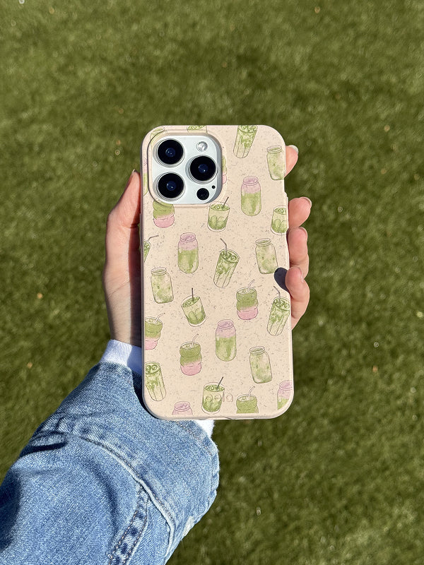 A hand holds a Pela phone case featuring the Matcha Love design. The case has a soft speckled beige background decorated with hand-drawn green and pink matcha drinks in various cups and jars. The phone has a triple-lens camera, and the eco-friendly case adds a fun, refreshing touch. The background is a sunlit grassy surface, and the person is wearing a light denim jacket.