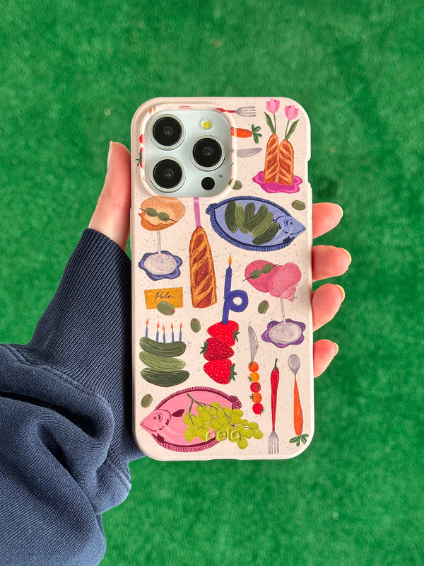 A hand holds a Pela phone case with a Surreal Soirée design, featuring a speckled beige background covered in vibrant illustrations of food and tableware—fish, fruits, bread, candles, and utensils—all arranged in a quirky, artistic style. The phone inside has a triple-lens camera, and the background is a bright green grass-like surface.





