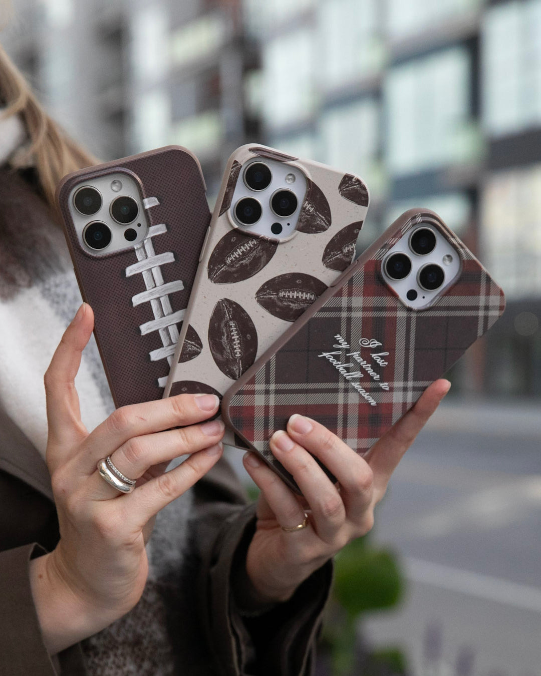 A woman holding all three Pela Touchdown Collection phone cases fanned out in both hands — featuring a close-up football lace design in dark chocolate brown, a tossed footballs pattern on Seashell, and the I Lost My Partner to Football Season tartan plaid in brown and red with white script text — with a blurred urban streetscape in the background.