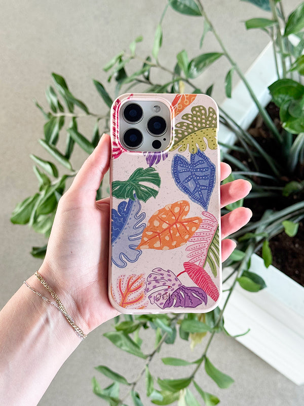 A hand holds a Pela phone case featuring the Tropical Cheers design. The case has a speckled light beige background decorated with colorful, illustrated tropical leaves in shades of green, orange, pink, purple, red, and blue. The phone has a triple-lens camera, and leafy plants in the background complement the vibrant, botanical theme. Made from Pela’s compostable material, the case combines eco-conscious design with bold tropical flair.