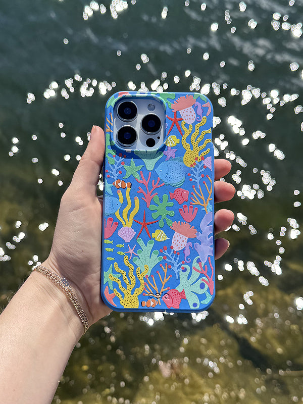 A hand holds a Pela phone case featuring the Underwater Blooms design. The case showcases a vibrant underwater scene with colorful coral, seaweed, starfish, seashells, and tropical fish in shades of blue, yellow, red, and pink. The background is a rich ocean blue, enhancing the marine theme. The phone has a triple-lens camera, and sunlight sparkles off the water in the background, adding a bright, beachy atmosphere. The case is made from Pela’s compostable material.