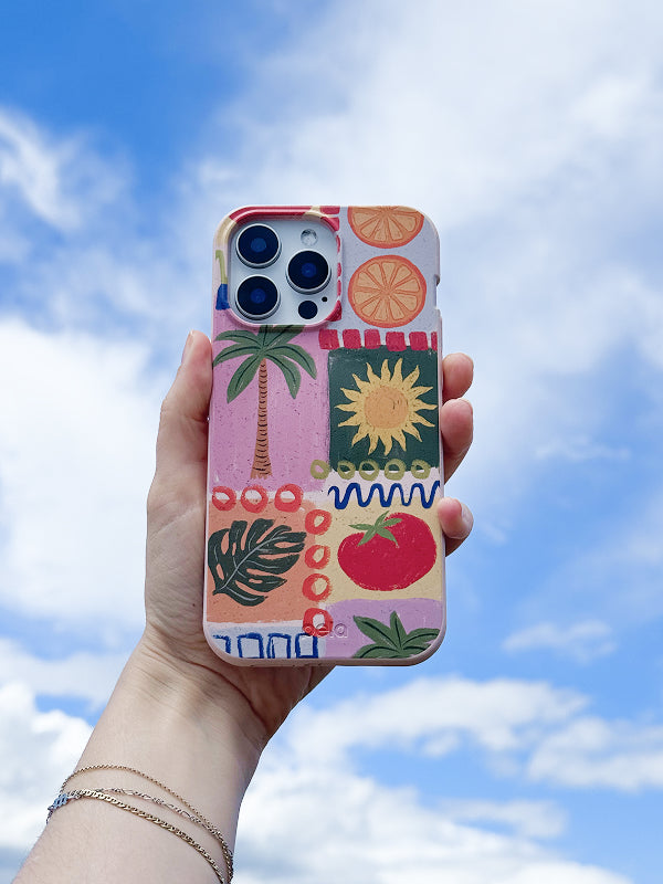 Hand holding a phone with the Hello Sunshine Case against a bright blue sky, featuring a colorful collage of tropical and summer-themed illustrations including a palm tree, sunflower, tomato, citrus slices, and abstract shapes.