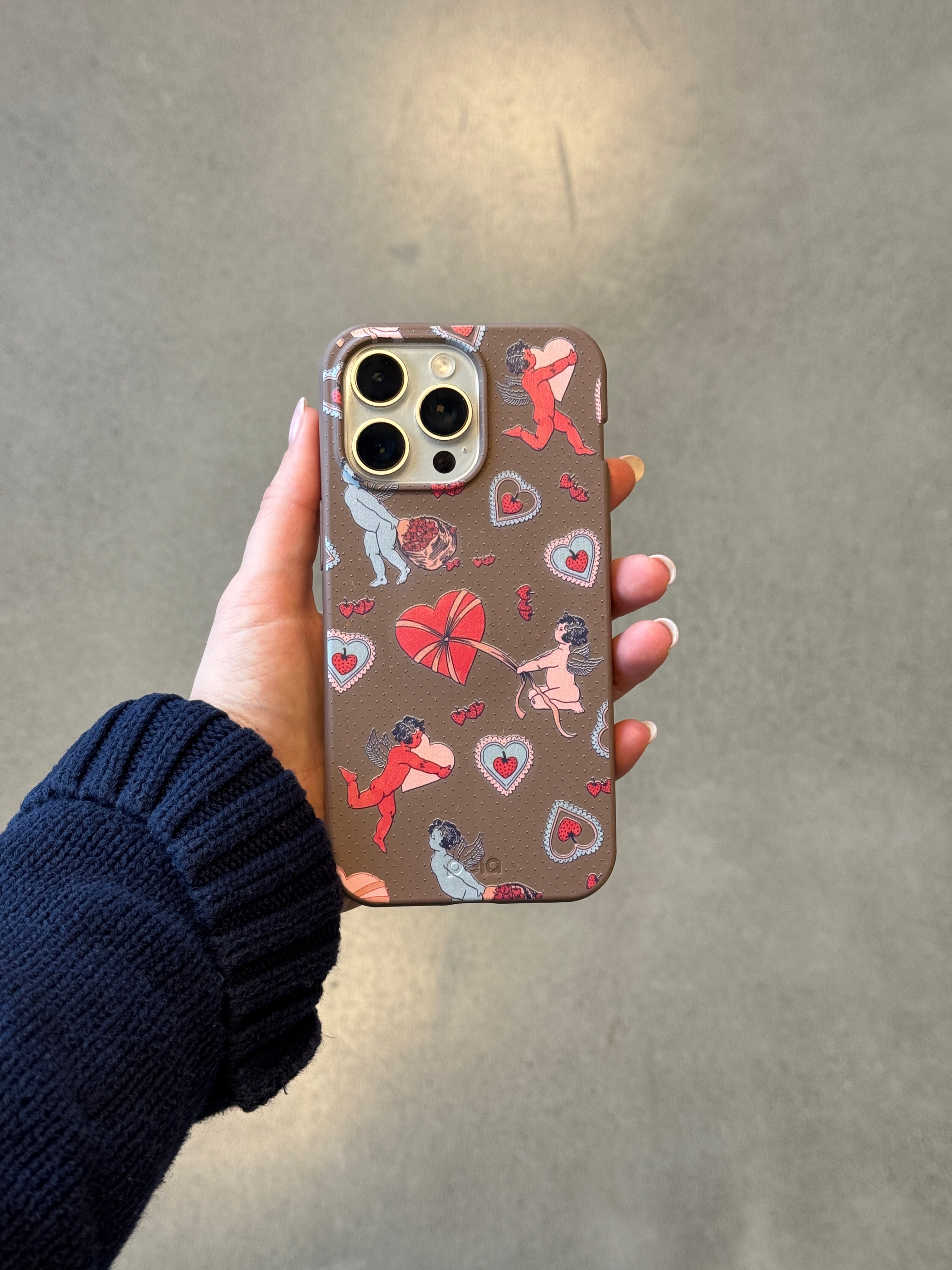 Hand holding iPhone with Pela's Strawberry Cupid phone case in chocolate brown featuring red and pink cupid cherubs with strawberry-colored curls, vintage lace hearts, and wrapped chocolates from the Valentine Collection.