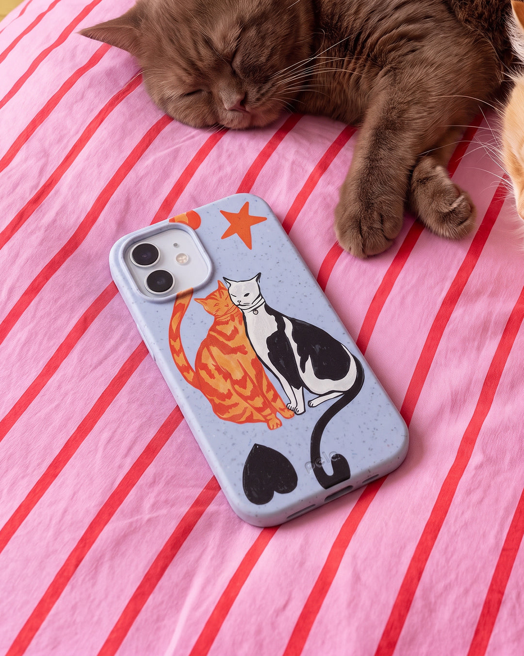 A Pela Twins phone case in Powder Blue featuring two illustrated cats — an orange tabby and a black and white cat — nuzzling together with a red star and black hearts, resting on a pink and red striped bedsheet beside a sleeping brown cat.